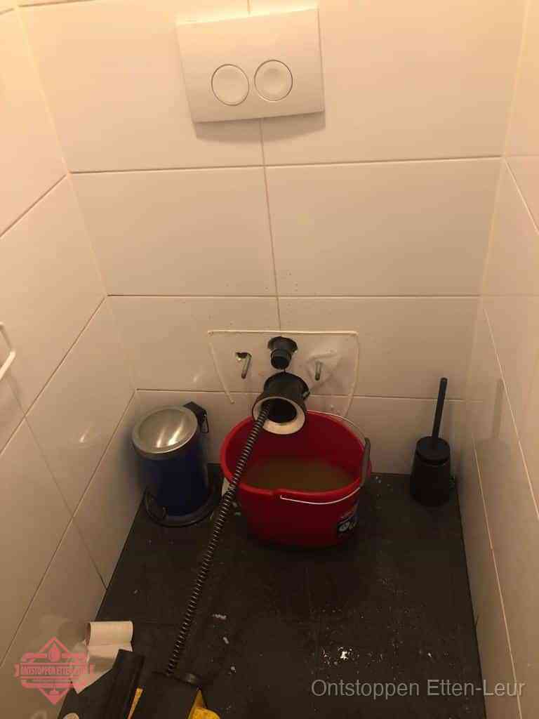 Een ontstopte wand-wc met verwijderde toiletpot waar een ontstoppingsveer in de wanduitgang verdwijnt, het achterhalen waar de verstopping zit in Etten-Leur