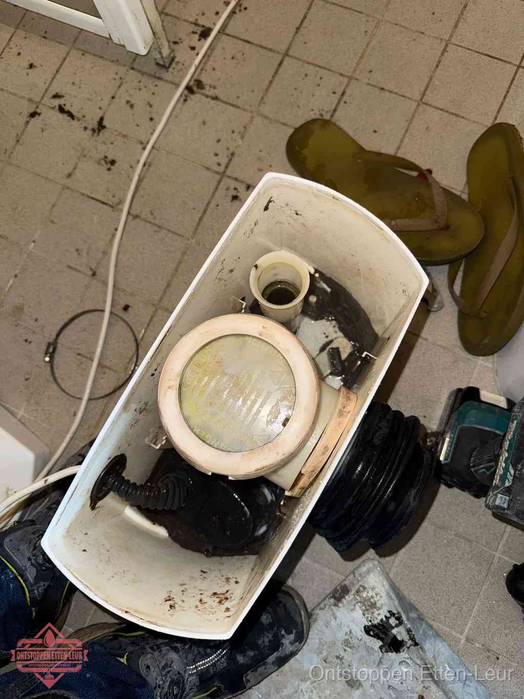 Een gedemonteerde toiletpot op de vloer met ontstoppingsveer en gereedschap ernaast, het verwijderen van een vastzittend voorwerp uit de wc in Etten-Leur